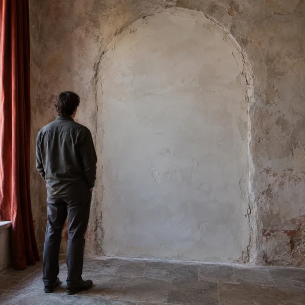 A traveler in dark clothing stands with their back to the camera before a sealed, plastered archway in a weathered corridor, with a rust-red curtain along the…