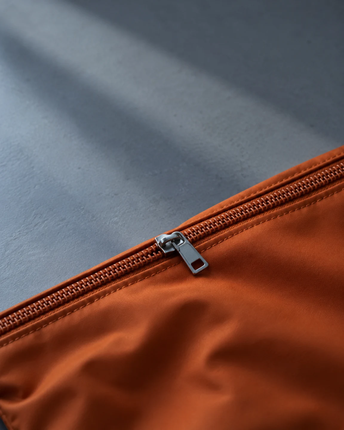 A burnt-orange fabric pouch fills the lower portion of the frame, with a closed diagonal zipper and silver slider against a cool grey tabletop.
