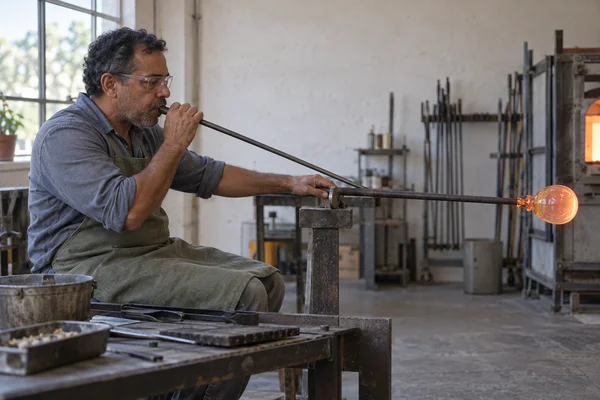 A middle-aged brown-skinned glassworker wearing clear protective glasses and a muted green apron blows into a long metal pipe while seated at a workshop bench.