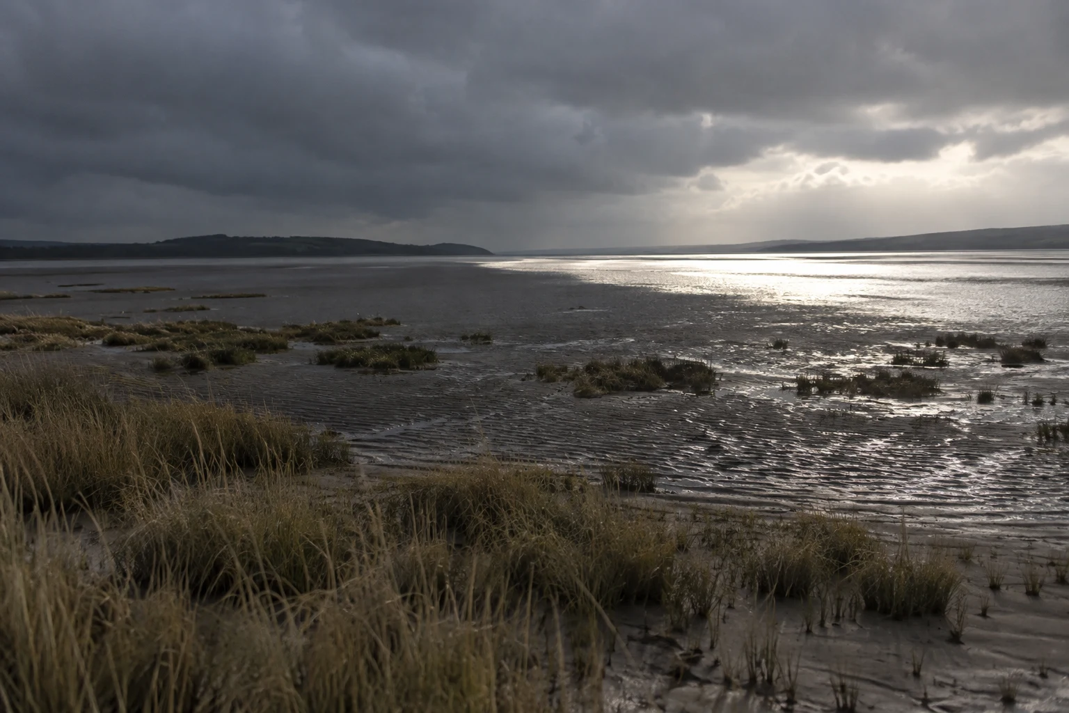 A wide estuary flat of rippled wet sand and scattered pale reeds beneath heavy grey clouds, with dark cloud shadow over the foreground and bright silvery tidal…