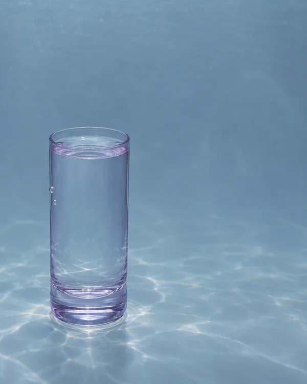 A pale lilac-tinted highball glass stands on a blue-grey floor in clear water, with two small bubbles on its outer wall and bright caustic patterns below and…