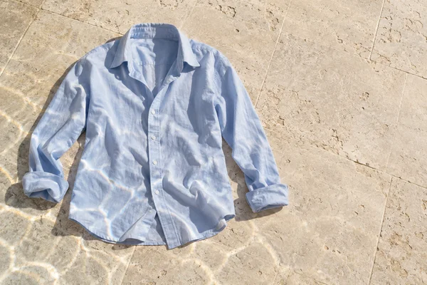 An overhead view of a pale blue button-up shirt lying on a warm ivory limestone terrace, with its sleeves loosely spread and bright wavering reflections…