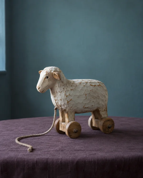 A worn white wooden sheep pull-toy with four wheels stands on a mauve linen-covered table, its rope string curling across the foreground against a muted teal…