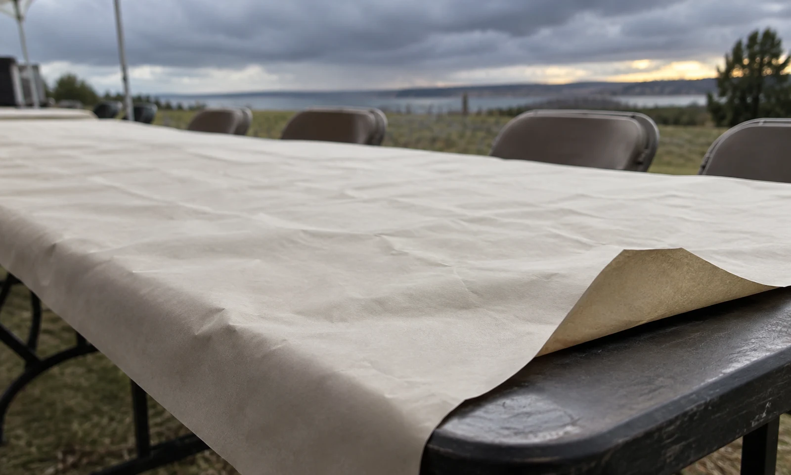 A broad sheet of unprinted cream paper covers an outdoor table, with a raised corner exposing the dark tabletop; empty folding chairs and a cloudy horizon sit…