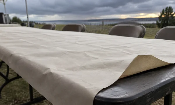 A broad sheet of unprinted cream paper covers an outdoor table, with a raised corner exposing the dark tabletop; empty folding chairs and a cloudy horizon sit…