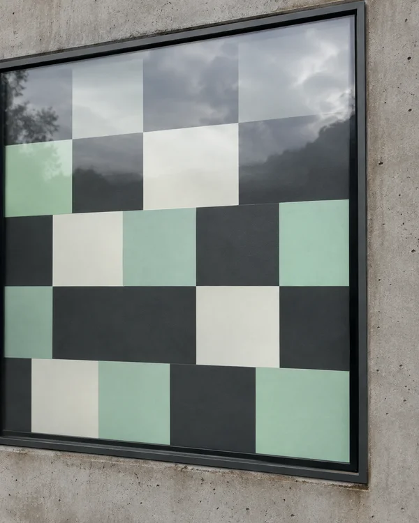 A large recessed digital display set into a rough concrete wall shows a mint, warm-white and charcoal checker pattern, with cloudy sky and tree reflections…