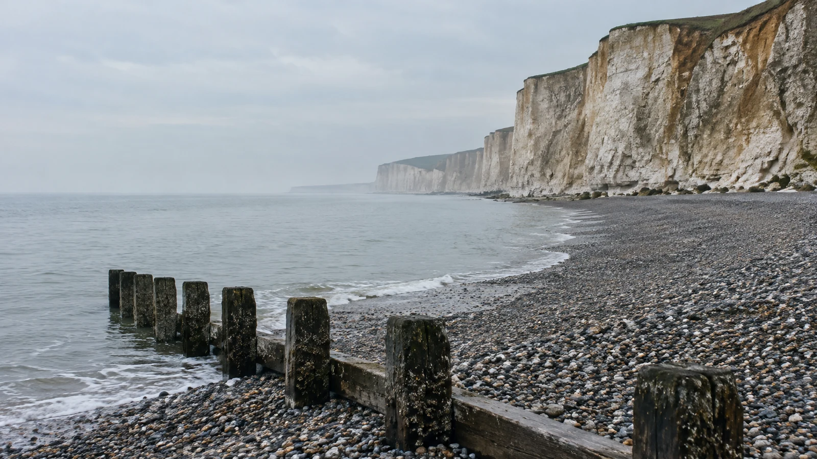 A weathered wooden groyne with dark posts crosses wet grey pebbles in the foreground of a quiet beach.