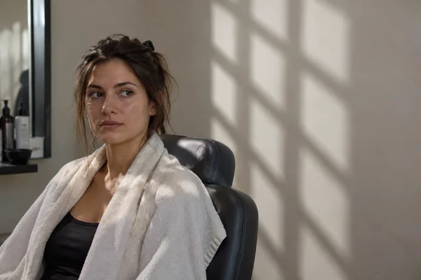 An adult sits in a black treatment chair in a beauty studio, wearing an ivory towel over a dark top with hair held back by a black clip.