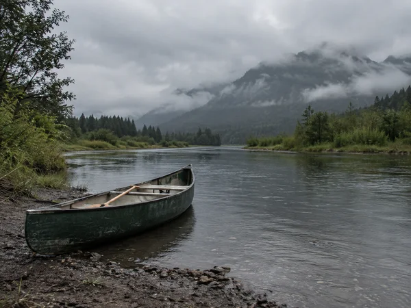 A weathered dark green canoe rests partly on a muddy gravel bank beside a broad river, with a paddle inside.