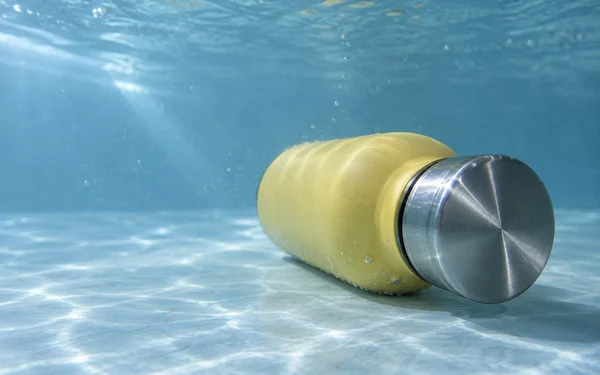 A matte butter-yellow bottle with a brushed-steel cap lies horizontally on a pale blue floor underwater, with the large circular cap facing the camera and open…