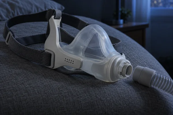 A translucent CPAP nasal mask with an ivory frame and charcoal woven headgear rests on dark gray upholstery; its hose connector is separated from the mask…