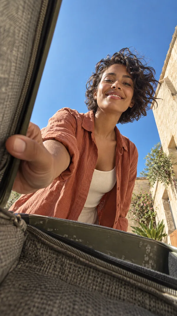 A smiling woman in a terracotta overshirt leans over an open olive suitcase in a sunny courtyard, with her foreshortened hand gripping its edge and blue sky…