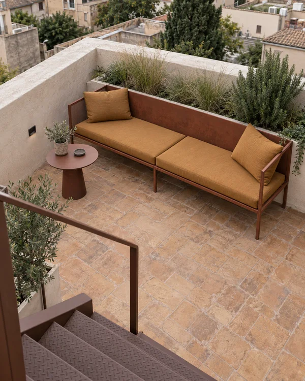 An elevated view of a residential terrace with a rust-coloured metal sofa and ochre cushions beside a round side table, surrounded by clay pavers and planted…