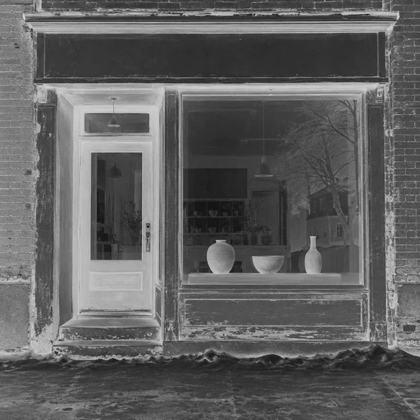 A weathered neighborhood storefront in a black-and-white negative treatment, with a recessed glazed door beside a display window holding three ceramic vessels.