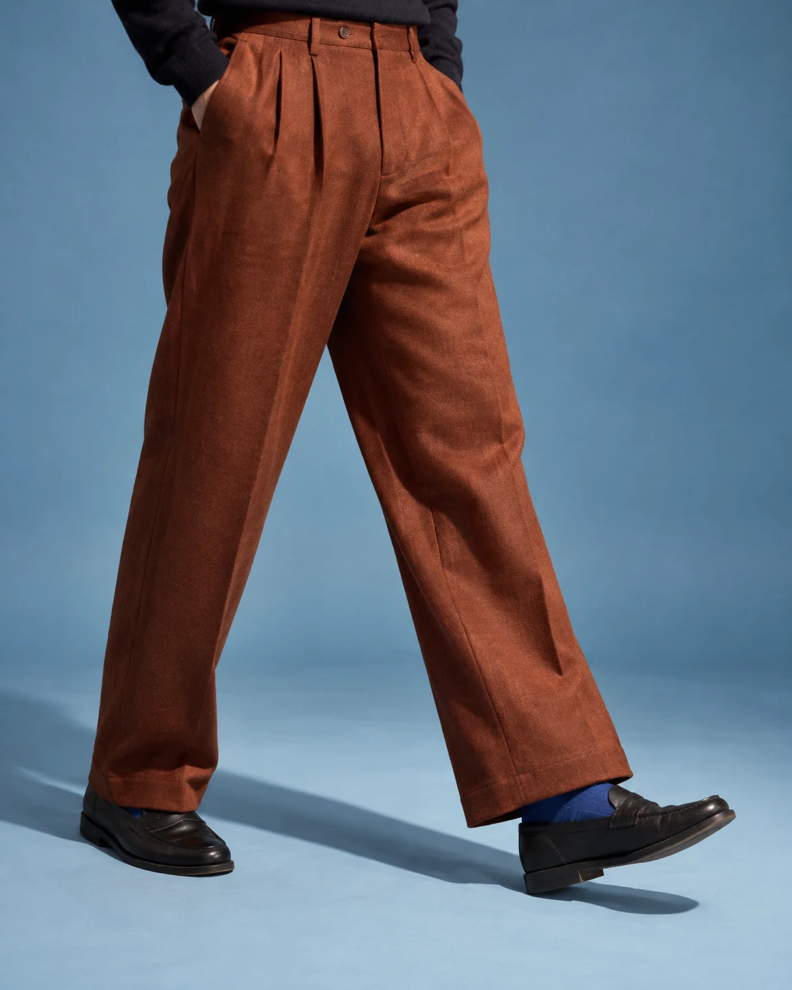 A person in rust-coloured pleated wool trousers, a dark sweater, cobalt-blue socks and black leather loafers takes a step across a pale blue studio floor, with…
