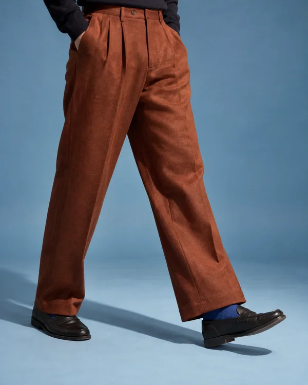 A person in rust-coloured pleated wool trousers, a dark sweater, cobalt-blue socks and black leather loafers takes a step across a pale blue studio floor, with…