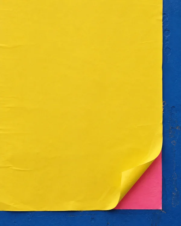 A large canary-yellow sheet covers a cobalt-blue posting board, with its lower-right corner curling up to reveal a hot-pink layer beneath; blue board borders…