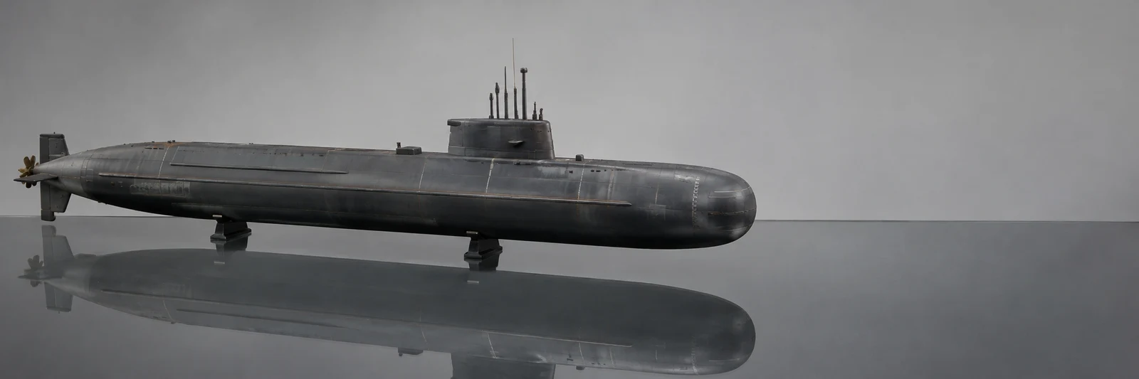 A long charcoal-grey submarine model rests on two low supports over a reflective glass tabletop, with its conning tower and propeller visible against a pale…