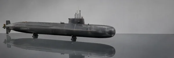 A long charcoal-grey submarine model rests on two low supports over a reflective glass tabletop, with its conning tower and propeller visible against a pale…