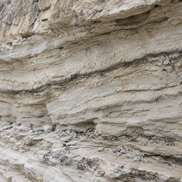 Close view of pale, weathered limestone with layered diagonal bands, a dark irregular seam crossing the rock, and small stone fragments on a ledge below.