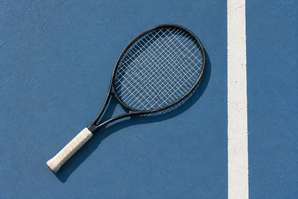 An unbranded dark tennis racket lies diagonally on a blue hard court beside a white boundary line, with its off-white grip pointing toward the lower left and a…