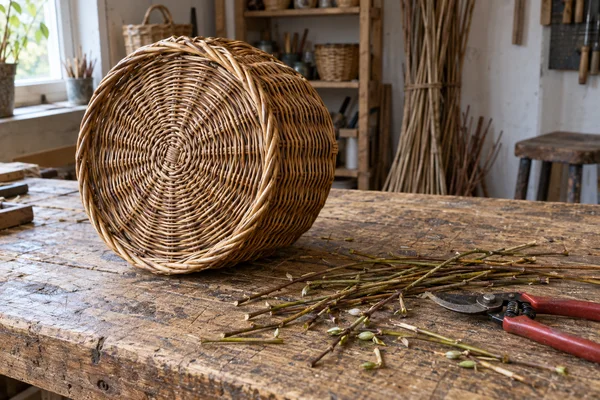 A round honey-brown willow basket rests on its side on a worn wooden workbench in a basket-making workshop, with its woven base facing the camera.