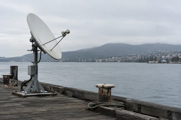 A weathered white satellite dish stands on a steel post at the edge of a timber wharf, with bollards and coiled rope in the foreground.