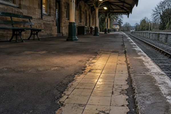 An empty heritage railway platform beneath a stone-and-iron canopy, with a worn strip of cream floor tiles exposed through dark asphalt beside the track.