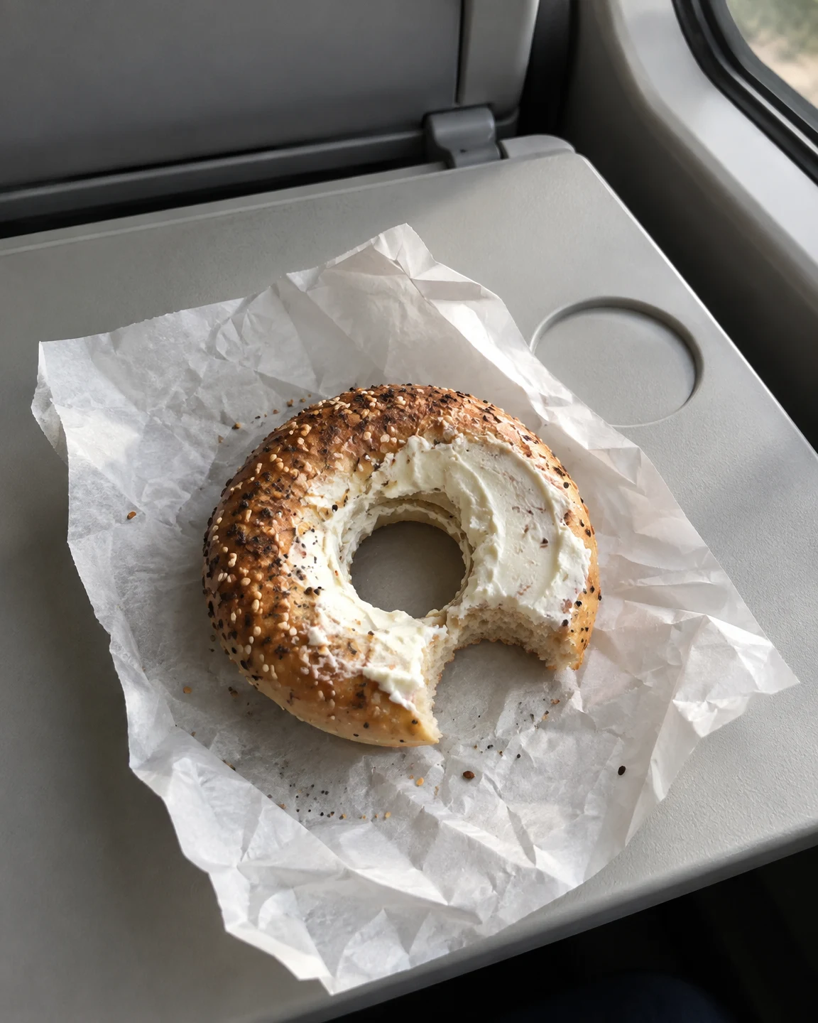 A cream-cheese bagel with a bite missing rests on a crumpled white paper wrapper on a pale grey tray, with a circular cup recess, hinge and window edge visible…