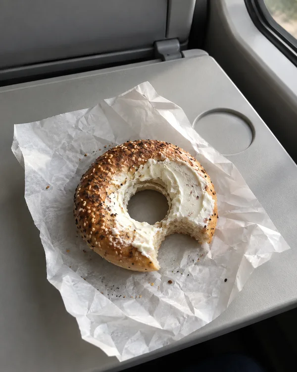 A cream-cheese bagel with a bite missing rests on a crumpled white paper wrapper on a pale grey tray, with a circular cup recess, hinge and window edge visible…