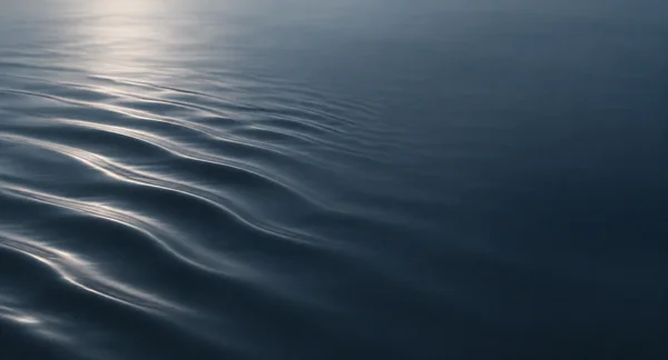 A blue-gray liquid surface fills the frame, with softly lit curved ripples sweeping in from the left and fading into a calm, dark area on the right.