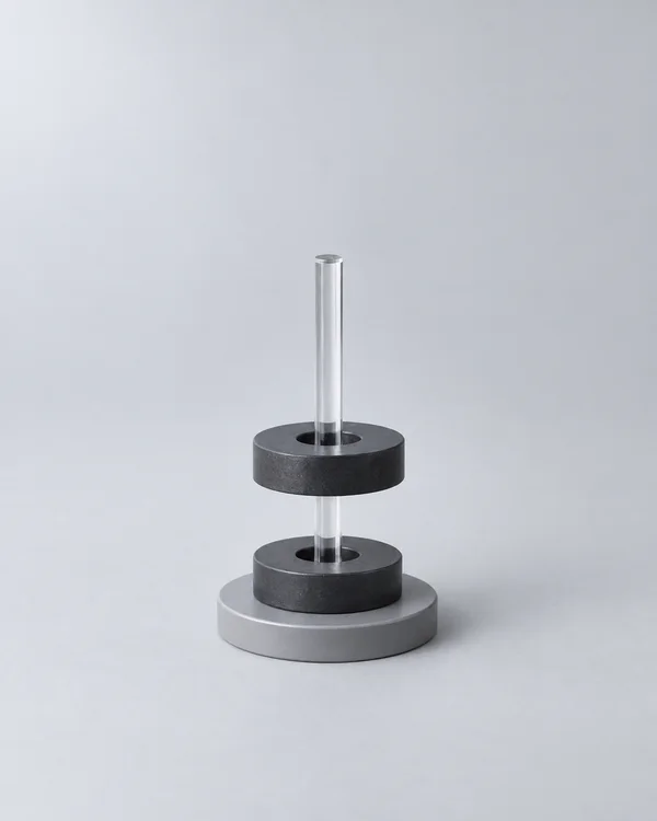 A clear acrylic rod passes through two charcoal ring magnets above a circular gray base on a pale-gray seamless surface; the upper magnet is visibly separated…