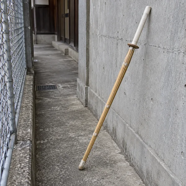 A bamboo kendo practice sword with a pale wrapped handle and round guard rests diagonally in a narrow concrete passage, its capped tip on the pavement beside a…