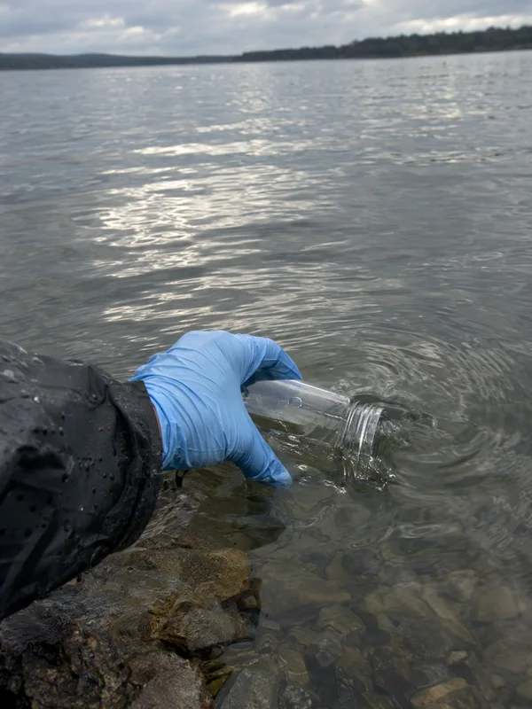 A blue-gloved hand extending from a dark waterproof sleeve holds a clear bottle partly submerged in a lake beside a rocky shore; a distant tree-lined bank is…