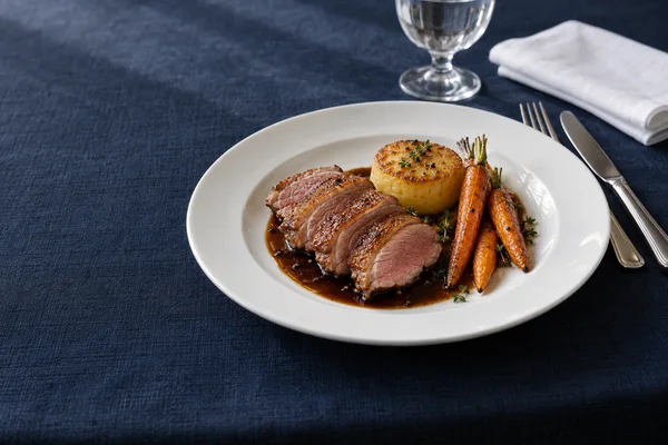 A sliced portion of rosy duck breast with crisp browned skin, a golden fondant potato and roasted carrots sits in glossy dark jus on a white porcelain plate…