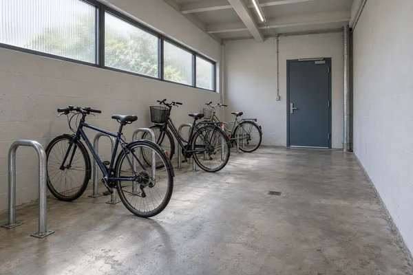Three commuter bicycles are parked beside metal U-shaped stands in a concrete-floored storage room with high windows, exposed ceiling beams and a closed…