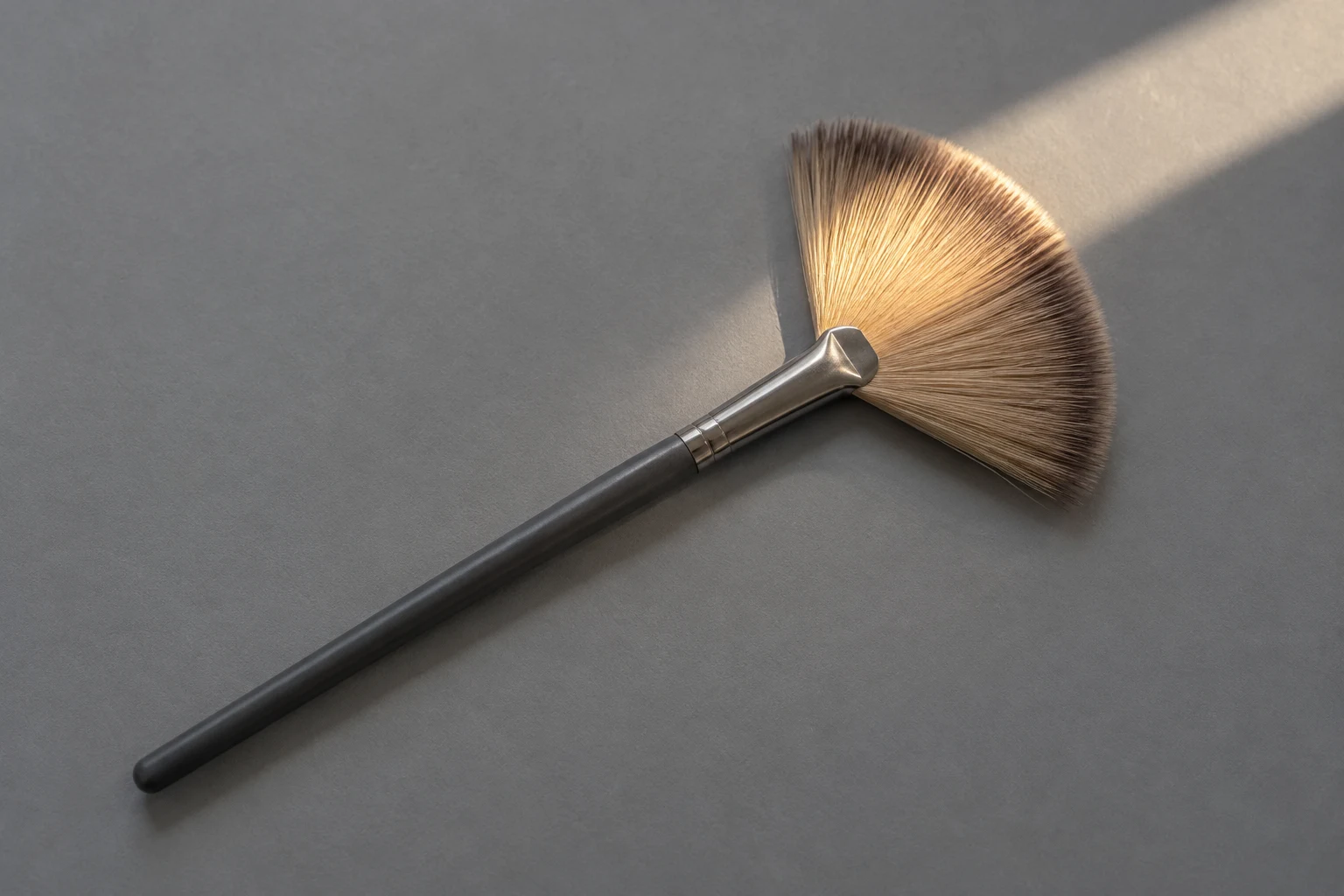 A single fan-shaped makeup brush lies diagonally on a cool-grey tabletop, with its charcoal handle pointing toward the lower left and tan bristles lit warmly…
