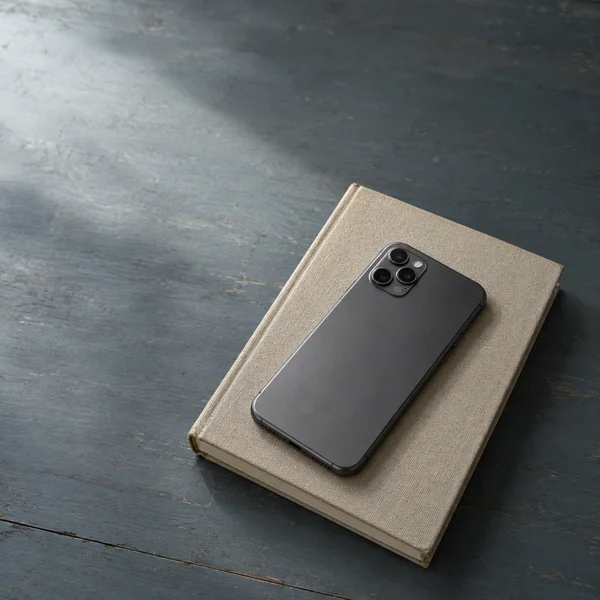 A graphite smartphone lies face down at an angle on a blank, oatmeal-colored clothbound book, placed on a worn blue-grey wooden tabletop.