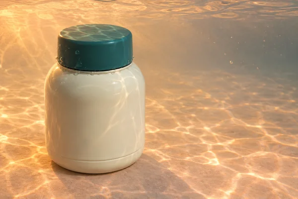 An upright ivory, rounded thermos with a broad deep-teal cap sits left of centre on a peach-toned tank floor, surrounded by clear water and warm orange light.
