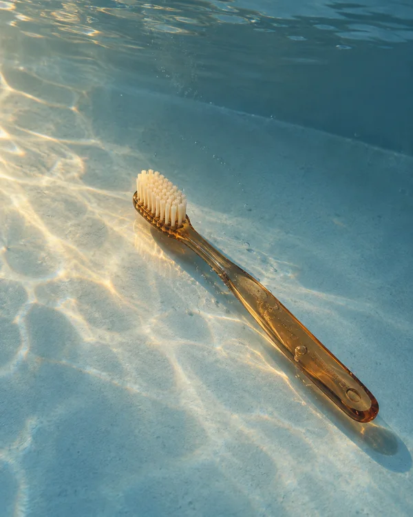 A translucent amber manual toothbrush with ivory bristles lies diagonally on a pale-blue submerged surface, lit by warm rippling highlights with small bubbles…