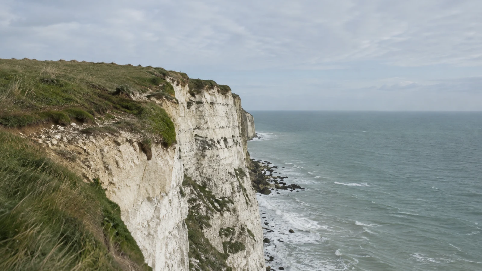 A grassy, windswept edge runs along pale, weathered chalk cliffs above a slate-grey sea with small waves; a softly clouded sky fills the background.