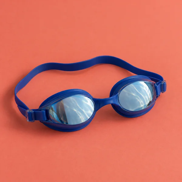 A pair of cobalt-blue swimming goggles lies on a coral-colored surface, with both reflective lenses facing upward and the strap forming a loose loop behind…