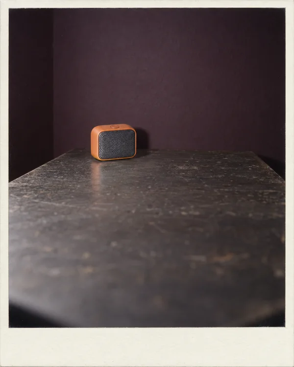 A small orange portable speaker with a dark perforated grille and one top button sits near the back of a heavily mottled charcoal workbench, against a…