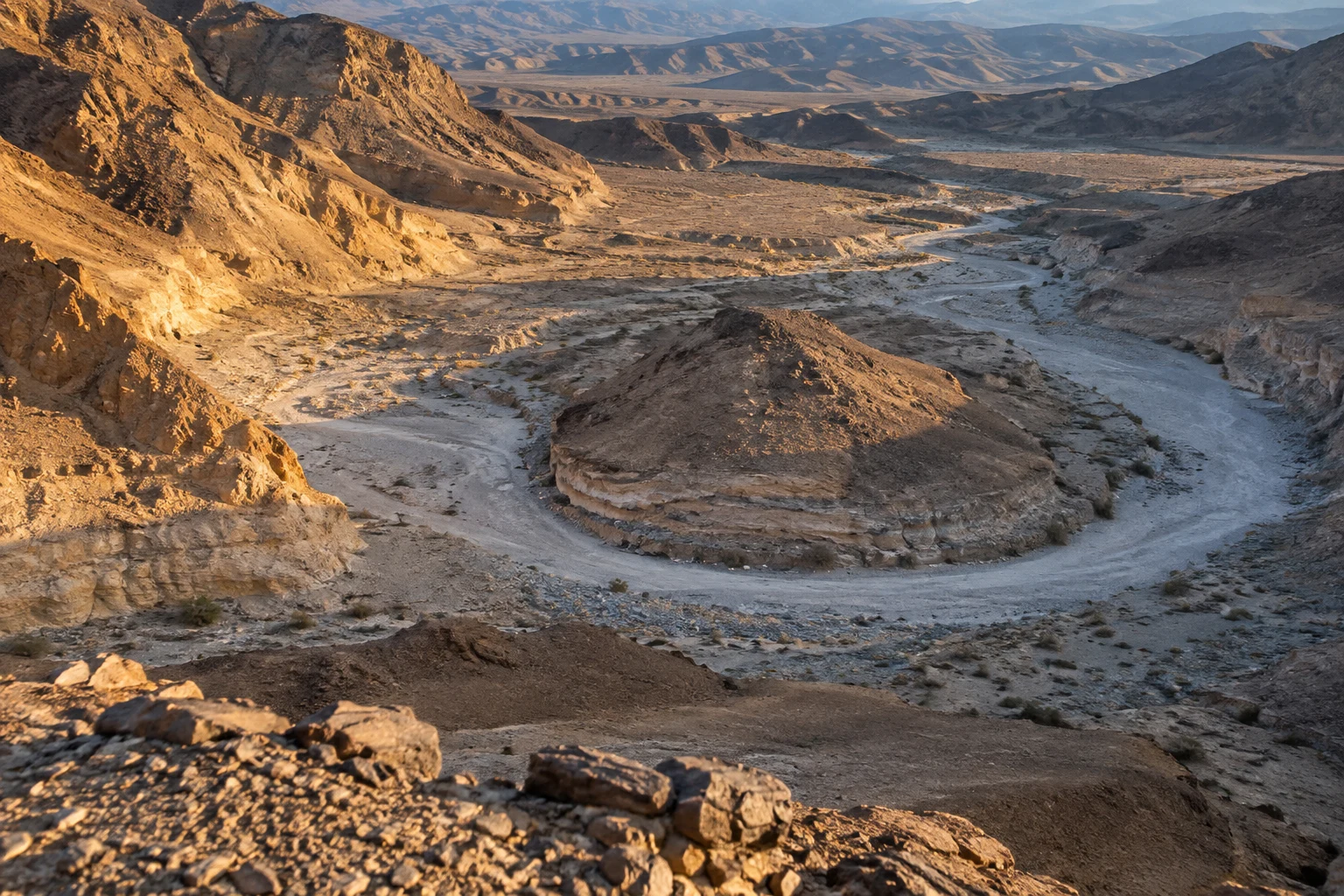 An elevated view looks down over an ochre desert valley, where a pale dry wash curves around a rounded, layered mound; warm sunlight strikes the left slopes…