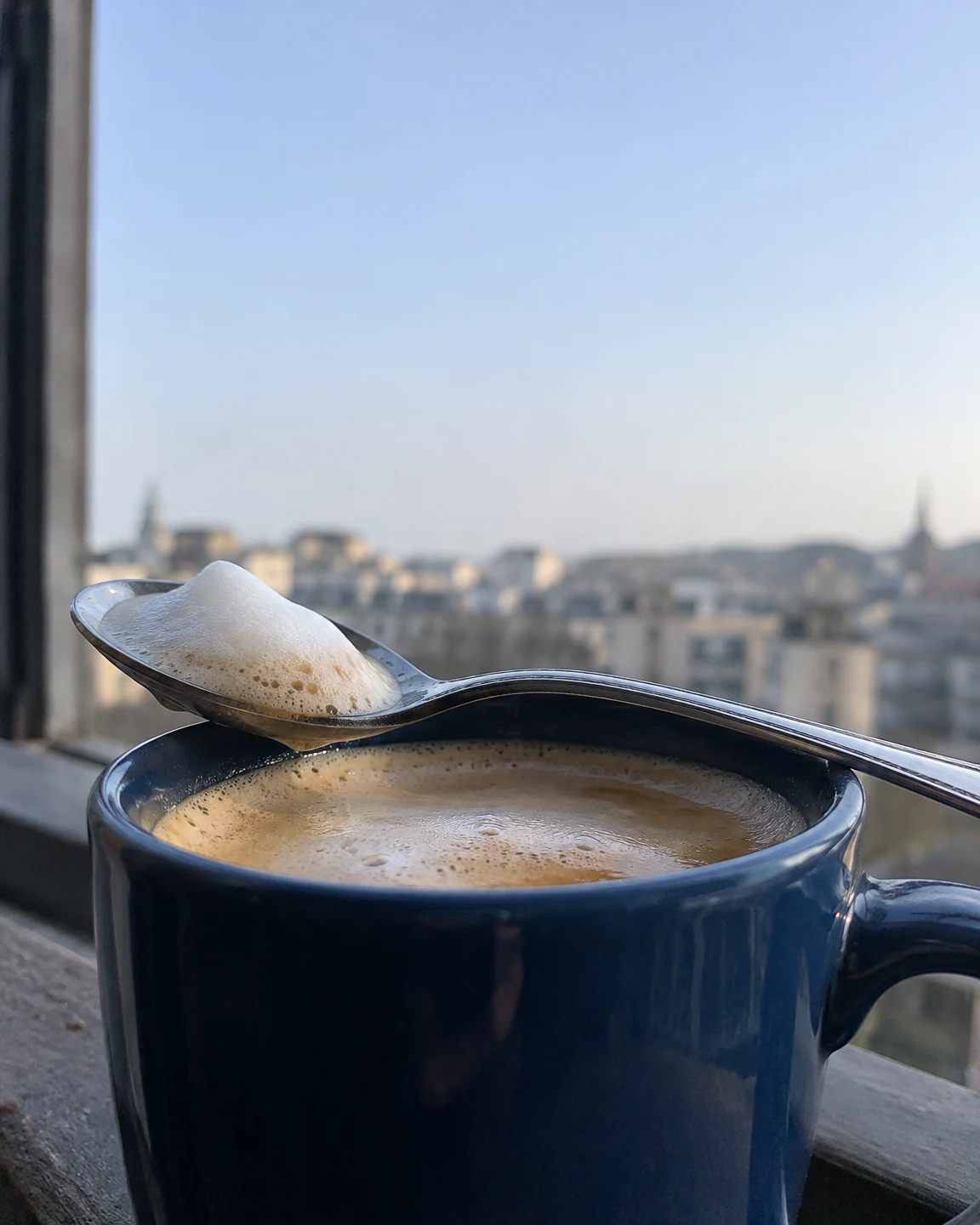 A dark-blue ceramic cup of coffee sits on a stone window ledge, with a stainless-steel spoon resting across its rim and a mound of pale foam in the bowl.