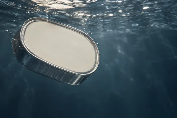 A sealed oval silver food tin with a blank ivory lid floats just beneath a rippled water surface, against deep blue water with open space to the right.