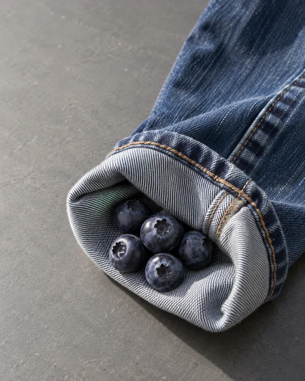 Five dark blue blueberries rest inside the turned-up cuff of faded indigo jeans on a grey tabletop.
