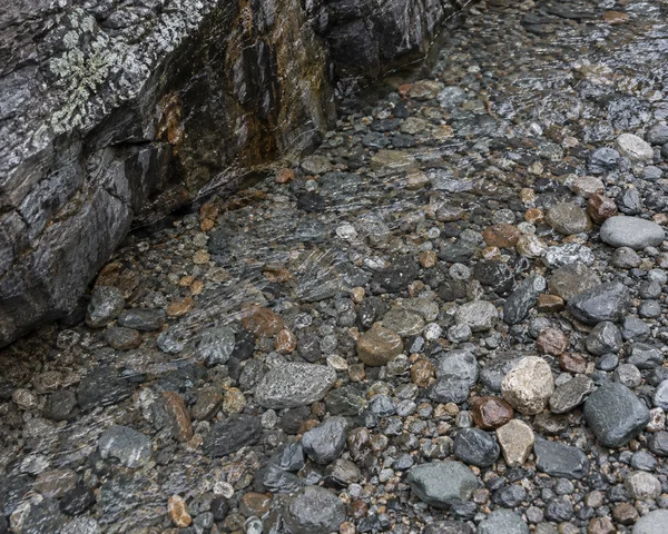 A steep downward view of tightly packed grey, rust and buff pebbles beside a dark, mineral-streaked rock face, with shallow water glinting in ripples across…