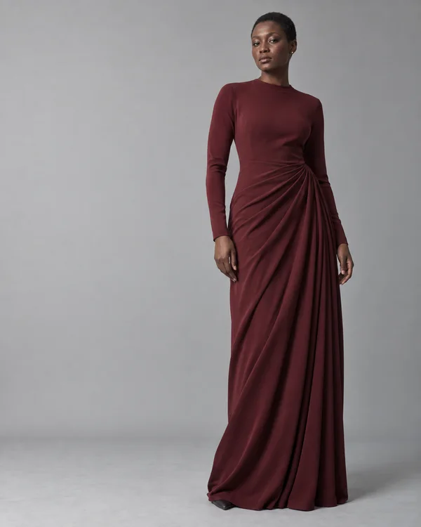 A full-length model with deep brown skin and closely cropped hair stands against a cool light-grey studio background, wearing a floor-length wine-coloured…