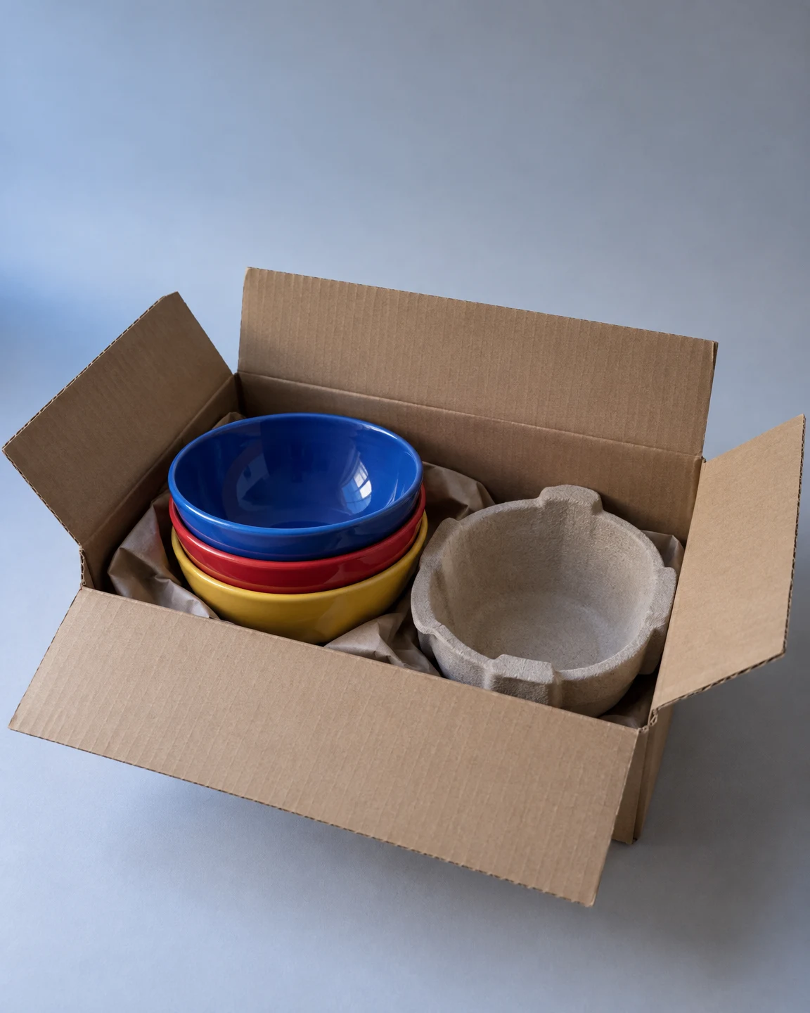 An open brown cardboard carton sits on a pale grey surface, holding three nested ceramic bowls with cobalt blue, red and yellow rims.